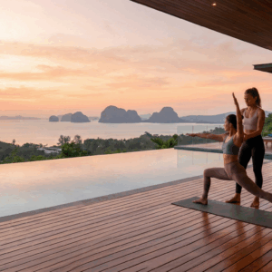 A woman practicing yoga with a private instructor on the wooden deck of a luxury villa in Phuket, overlooking the ocean at sunrise.
