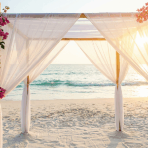 A romantic bamboo wedding arch draped in white fabric and tropical flowers on a sandy Phuket beach during sunset.